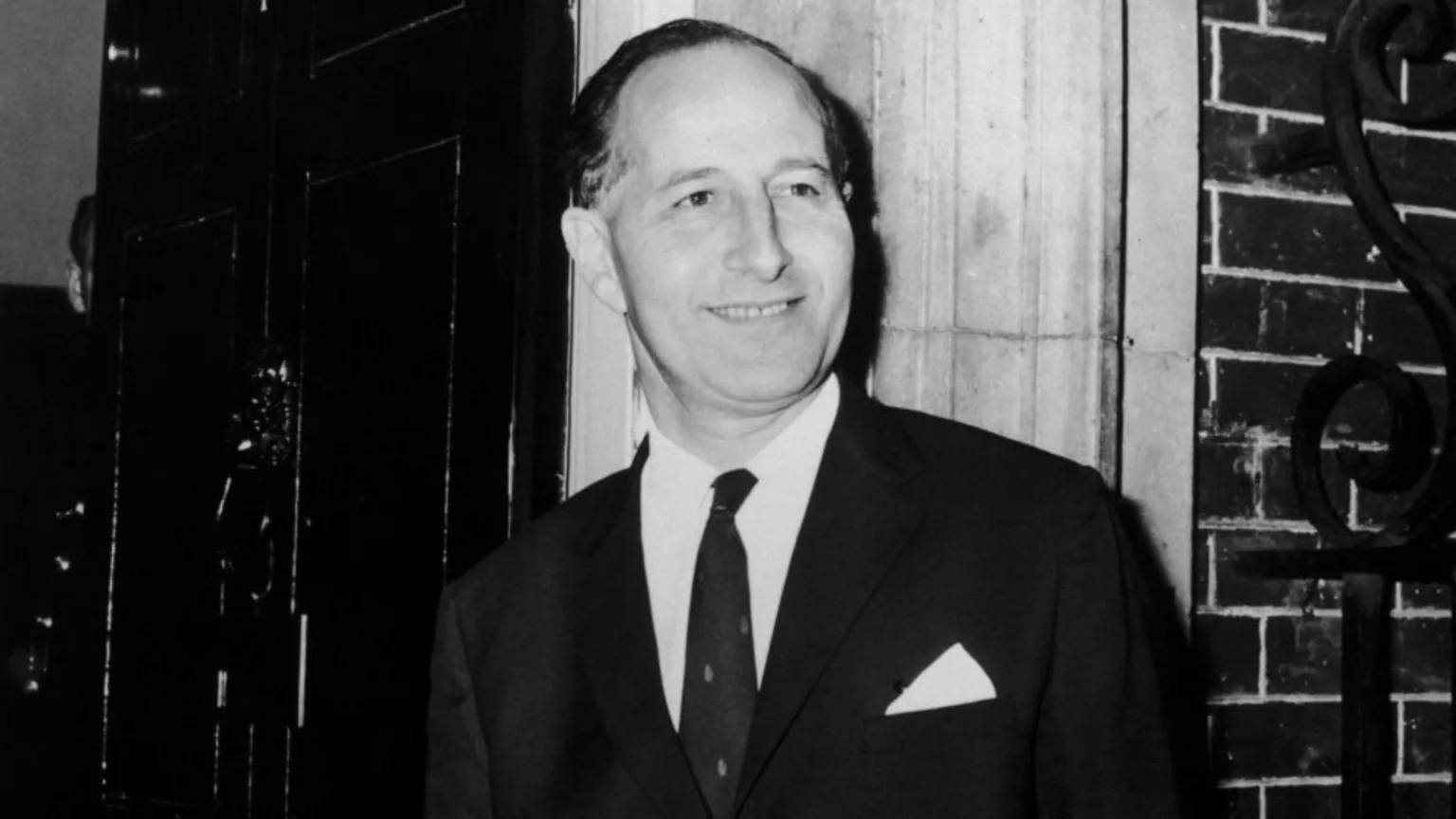 Black and white photograph of Terence O'Neill, he has short hair and is smiling to the side. Wearing a suit jacket, shirt and tie. Pictured outside a bricked building.