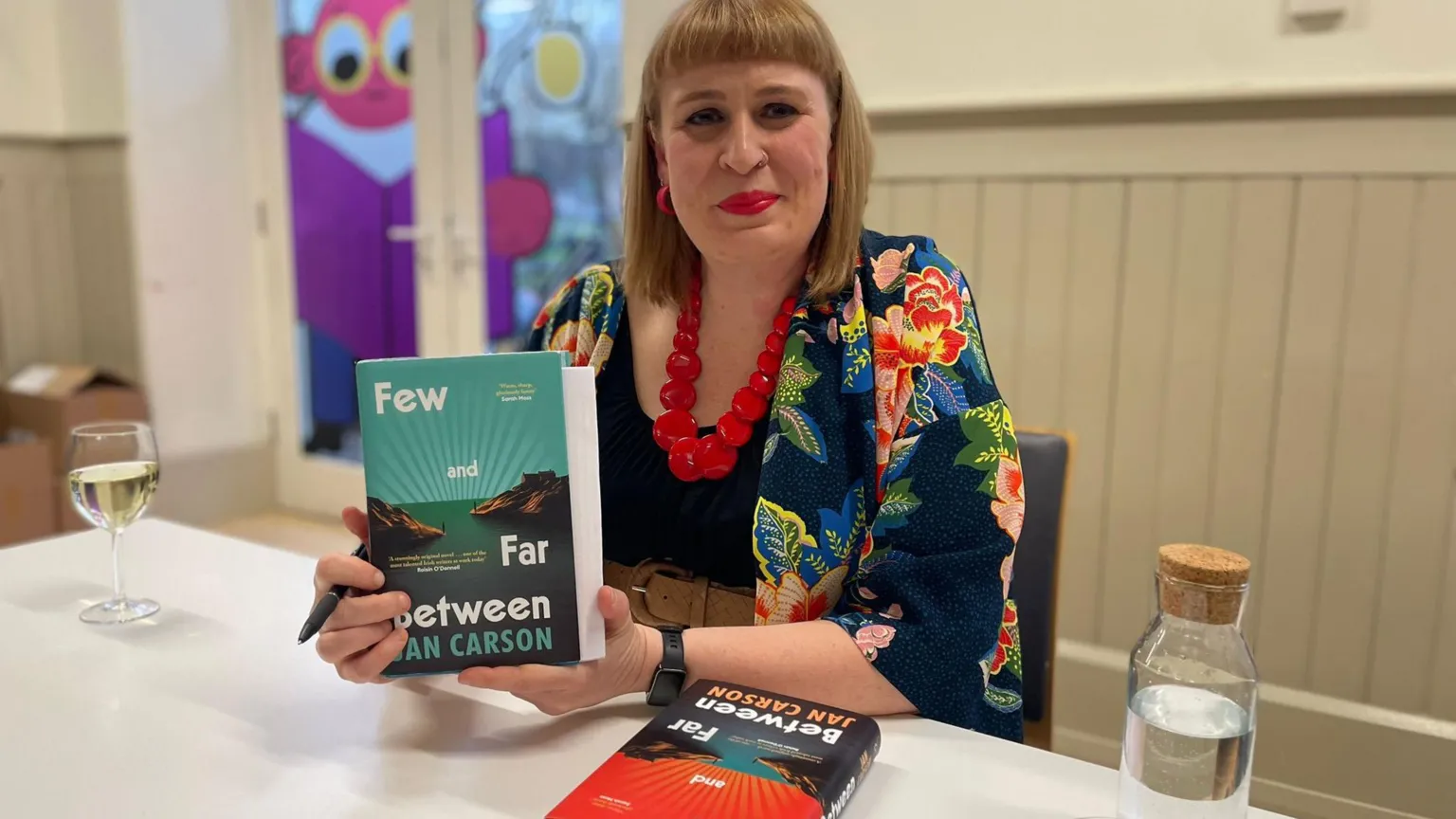 Jan Carson poses with a copy of her book Few and Far Between. She is wearing a black coat with flowers on it