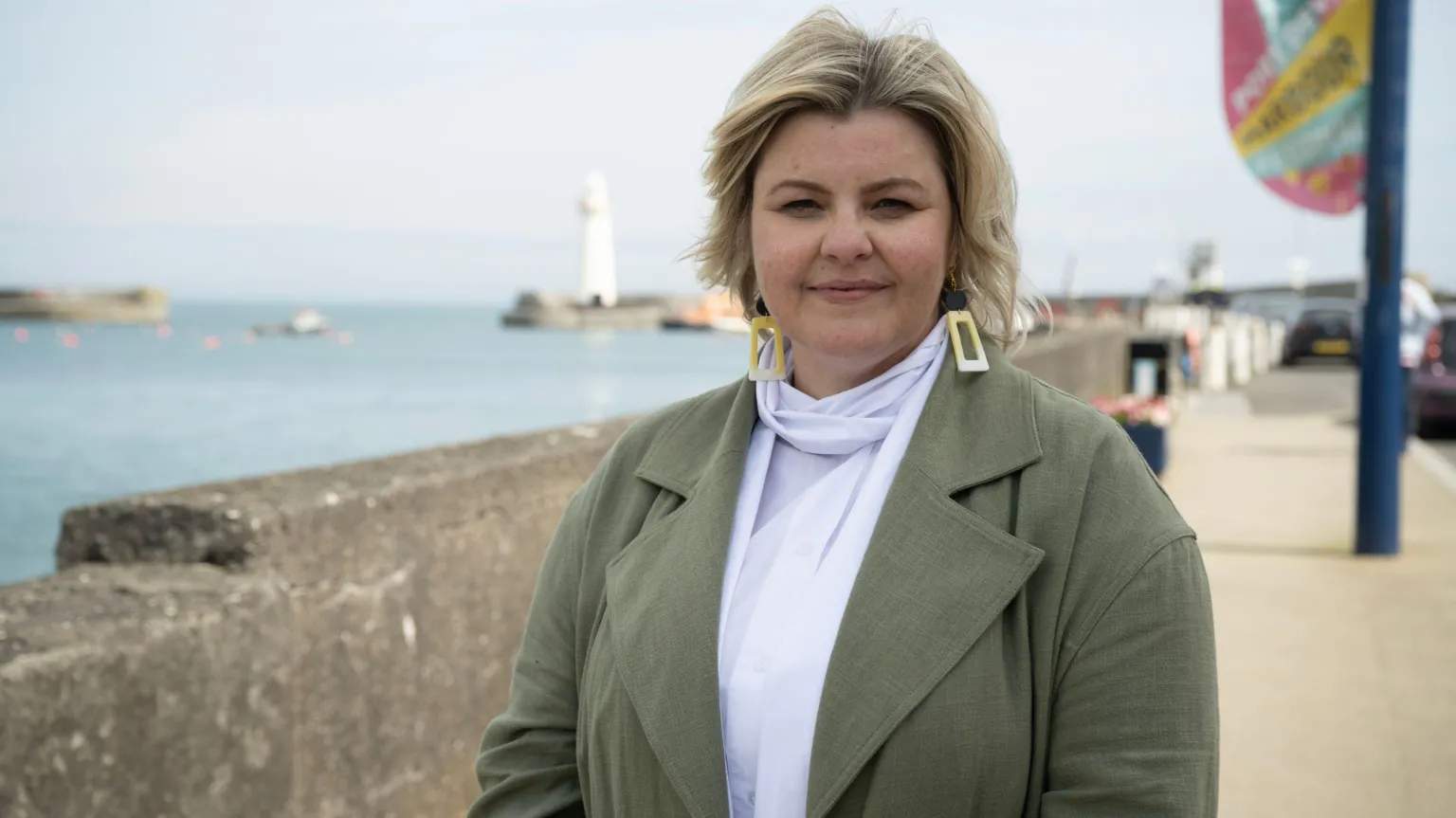 BBC/Long Story TV/Jack Maguire A woman standing at a pier, she has a green coat and a white blouse on. She has blonde hair.