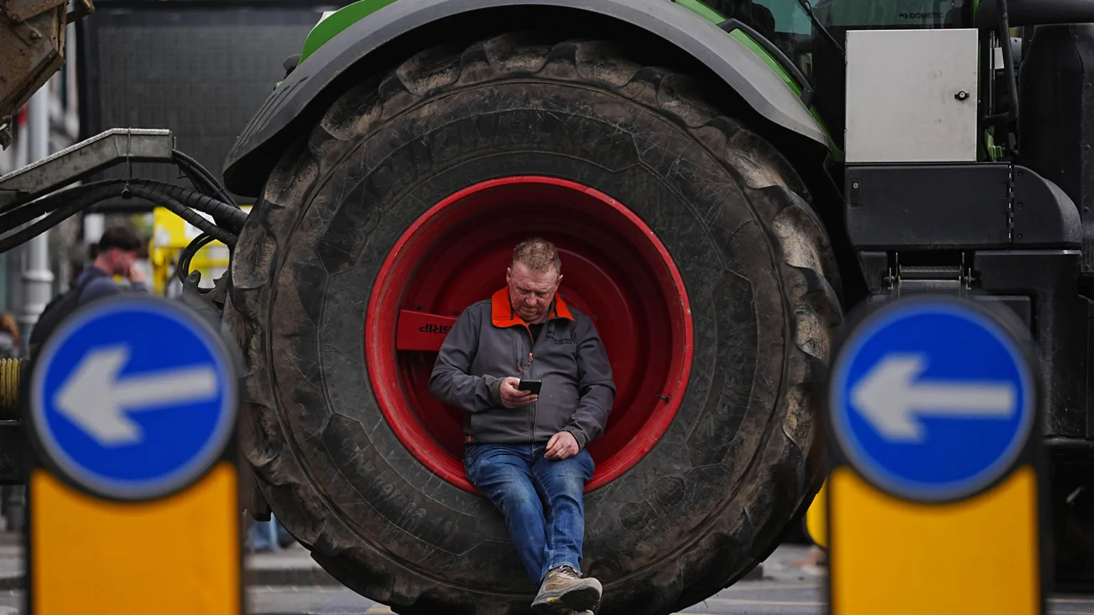 Brian Lawless/PA Wire A man sits inside a large tractor wheel as vehicles are parked on O'Connell Street in Dublin. He's looking at his mobile phone. He's wearing a navy jacket with a red collar, blue jeans and brown boots.