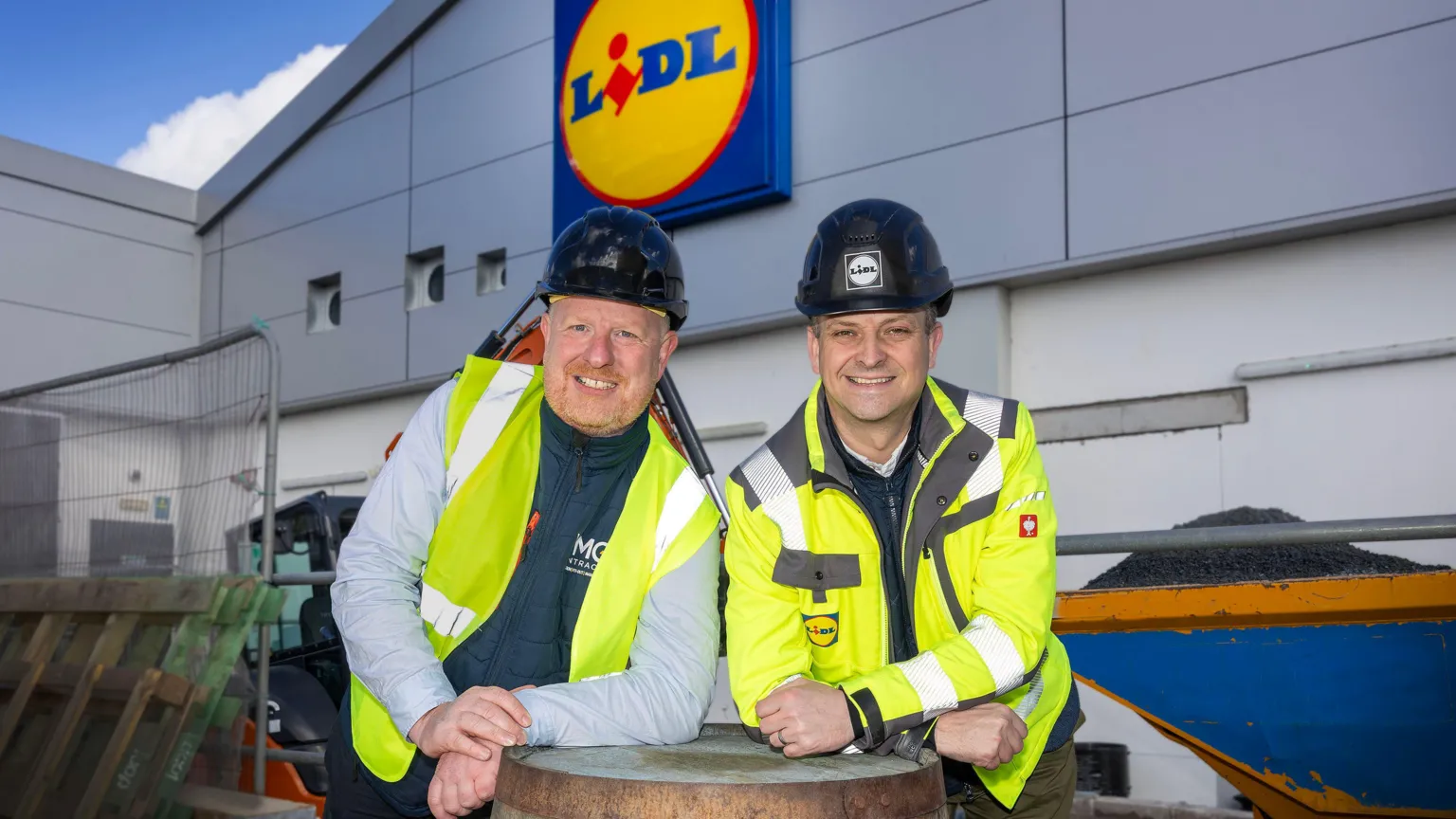 Lidl Two men with black hard hats, high vis jackets leaning on a barrell. The Lidl logo is behind them