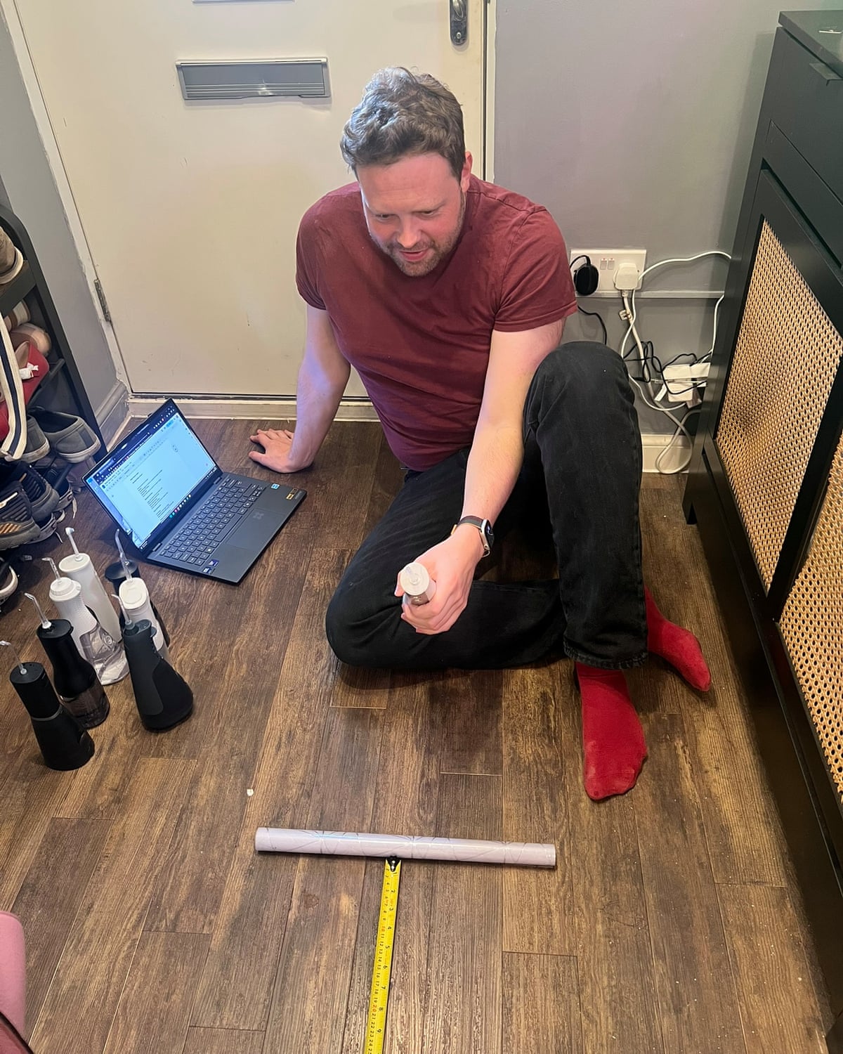 A man sits on a wooden floor holding a water flosser with a tape measure in front of him 