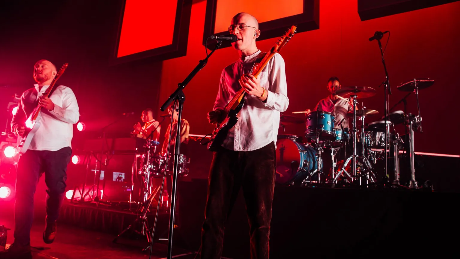 Bombay Bicycle Club play on stage and there is a red hue over the stage. There is a guitarist playing and singing into a microphone in the middle of the stage.
