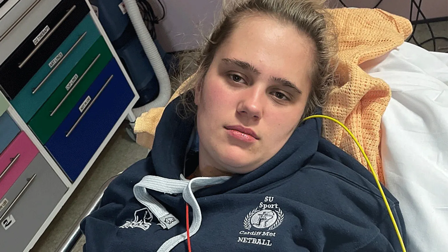 Phoebe Tesoriere Phoebe led in a hosptial bed wearing a blue Cardiff Met netball hoodie.