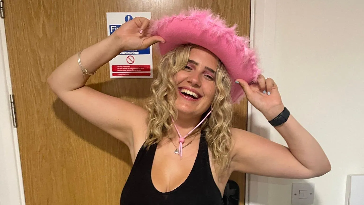 Phoebe Tesoriere Phoebe who has mid length blonde curly hair is posing in front of a light brown wooden door. She is smiling whilst wearing a pink cowboy hat with a feathered trim. She is holding the cowboy hat on her head with both hands and her arms raised.