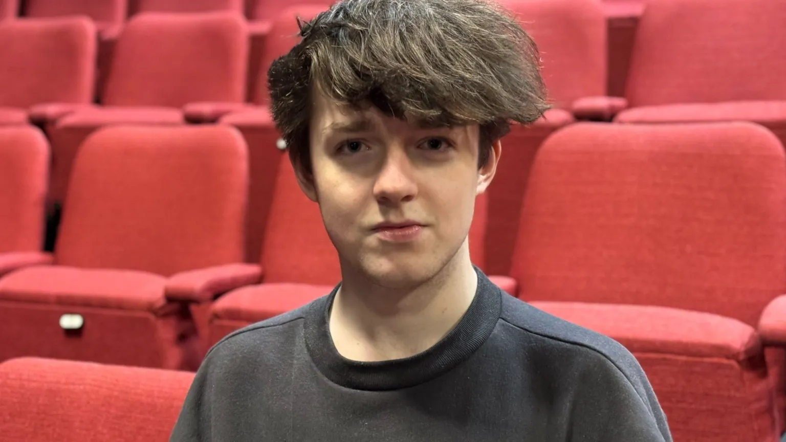 Boy with brown hair in front of red seats wearing a black top.