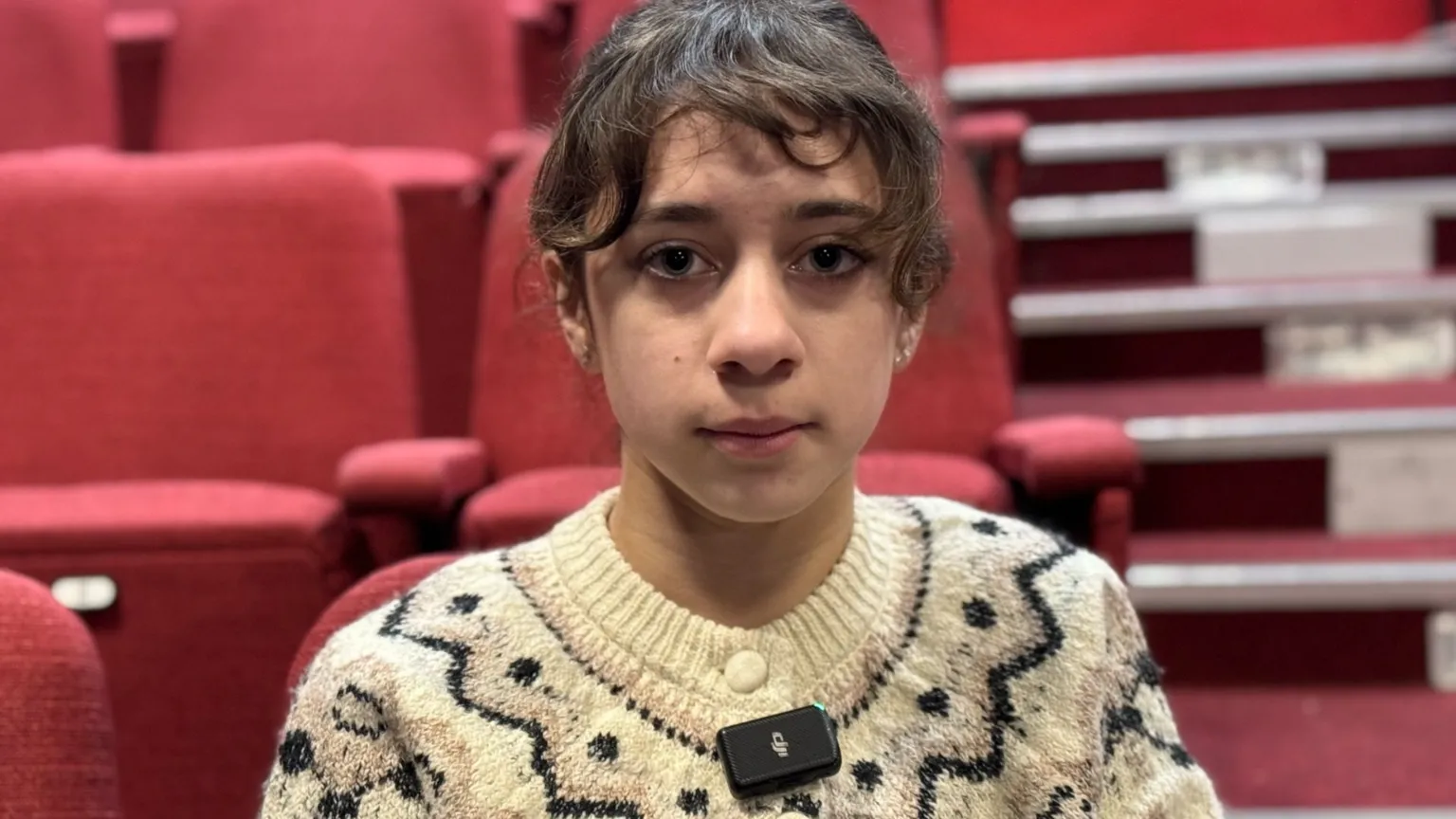 Girl in white patterned jumper in front of red seats.