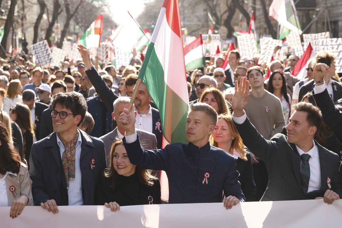 Magyar, front centre, during a rally in Budapest. People hold flags behind him with some raising their hands in the air. It is sunny in a wide, tree-lined street and the people around Magyar look happy and smiling. 