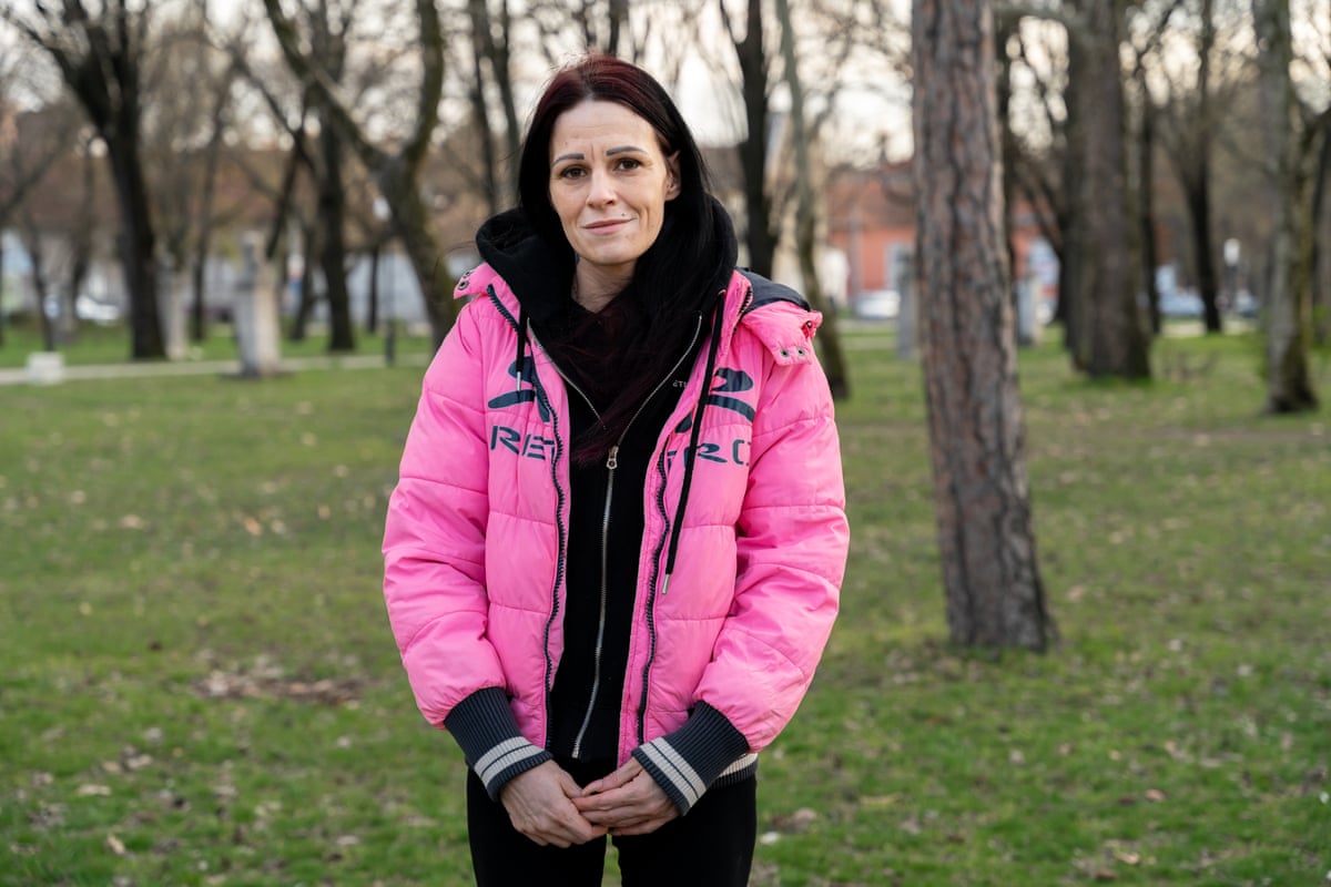Anita, 33, stands among small trees in a park. She has long, straight dark hair and very pale skin, and wears a black hooded top under a bright pink padded anorak. 