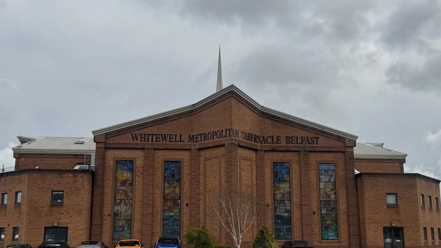 The exterior of Whitewell Metropolitan Tabernacle in Belfast, a large red‑brick building with stained‑glass windows and a central spire, photographed under a cloudy sky.