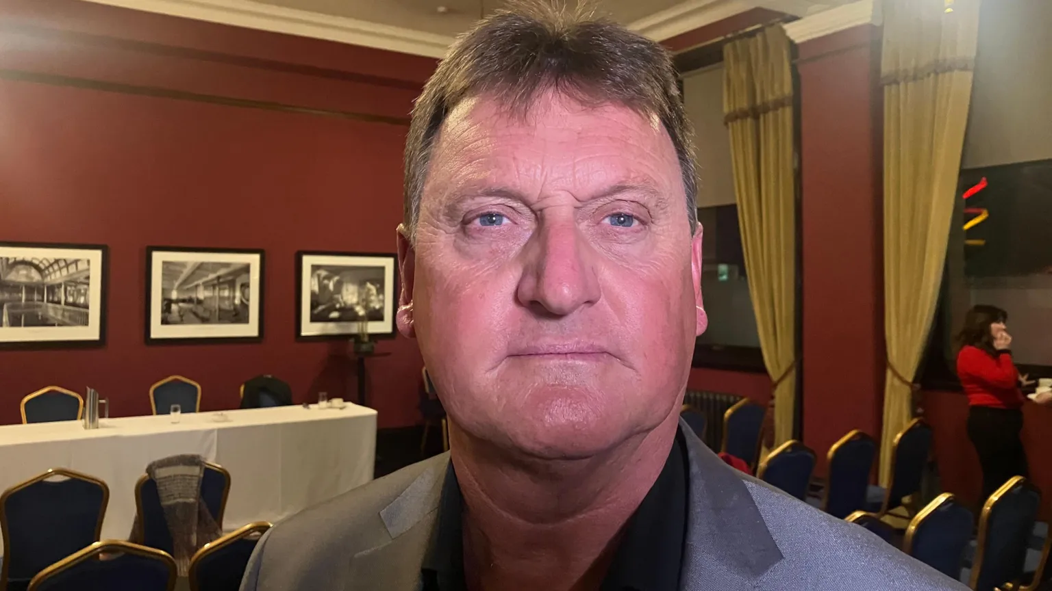 A man with dark hair, wearing a black shirt and a grey jacket