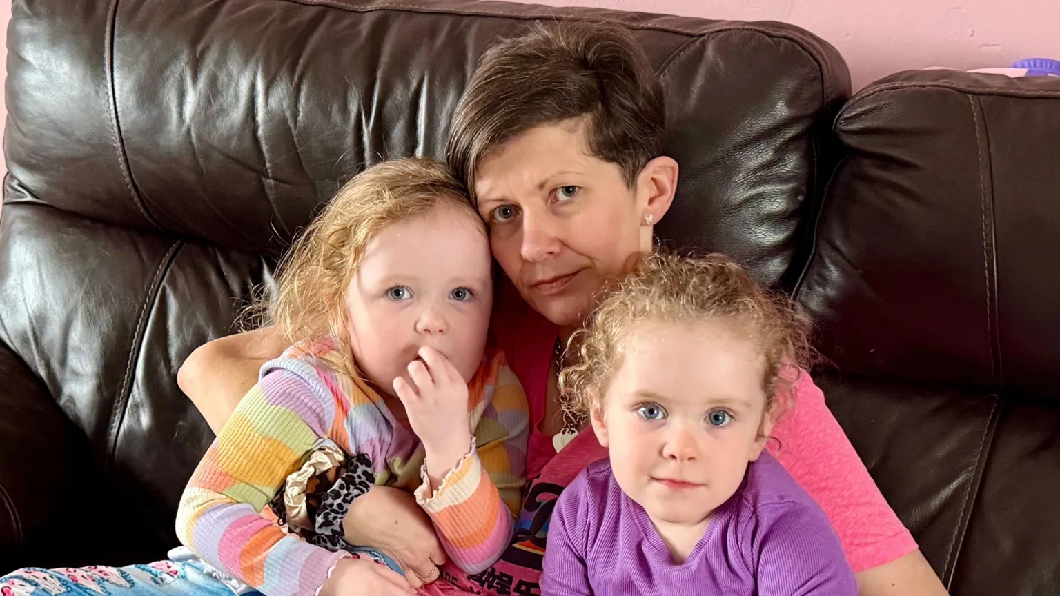 Linda McBride A woman with short, brown hair sits on a dark leather sofa with two young children on her lap, indoors. The woman wears a pink top, while the children wear colourful jumpers and jeans, looking towards the camera.