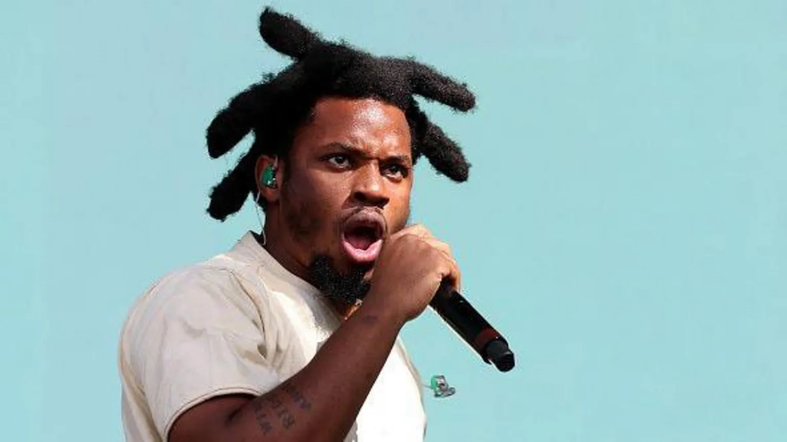  Denzel Curry has thick black hair styled on each side of his head. He wears a white t-shirt, a green ear-piece and holds a black microphone as he performs against a turquoise background. He has a tattoo on his forearm.
