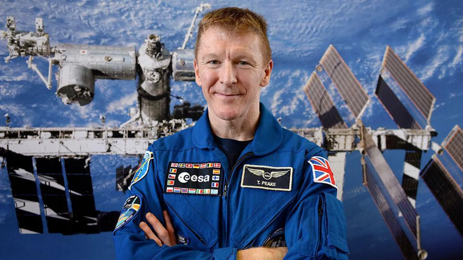  Tim Peake, a man, wearing blue space overalls and crossing his arms, smiling. Behind him is an image of the International Space Station.