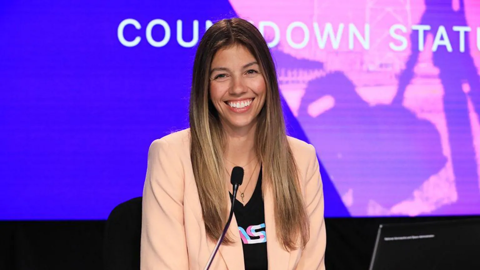 NASA A woman wearing a cream coloured blazer, smiling. There is a black microphone in front of her, with an LED background of blue and purple, with text in white that reads 