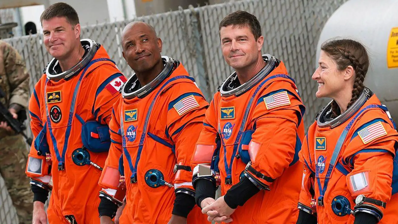 Four astronauts wearing orange Nasa spacesuits, there are three men and one woman on the right. She has brown hair in two plaits. Both men on the far left and second from the right have short brown hair. The second man is bald. 