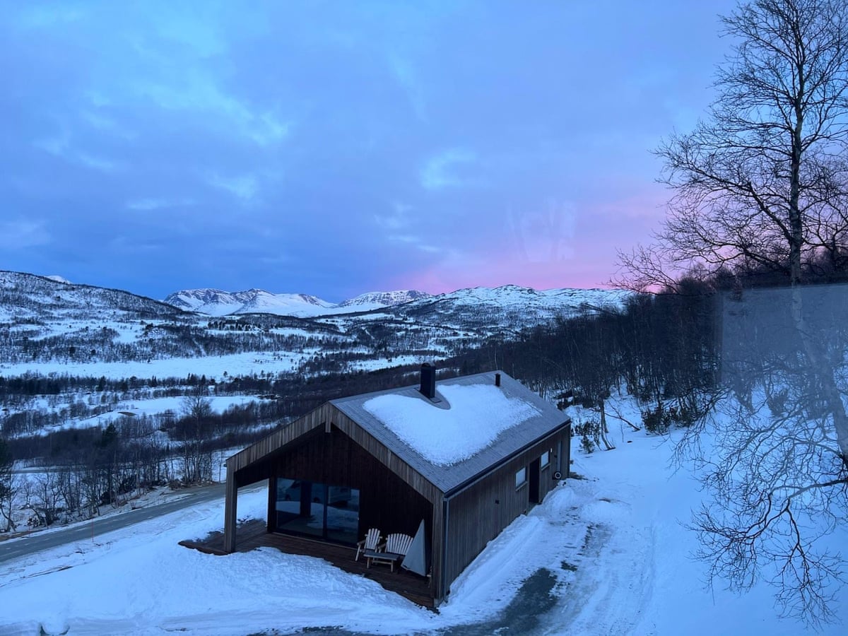 A snow-laden chalet in Norway overlooking a mountain range; in the distance, bare trees and an evening sky with clouds turning pink at the horizon