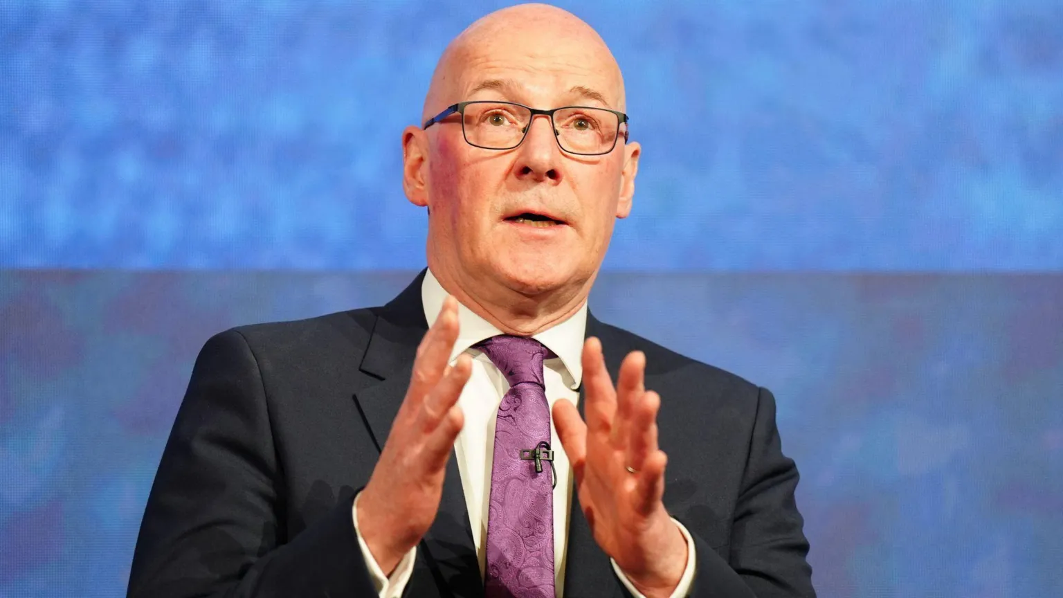 John Swinney wearing a dark suit over a white shirt and purple tie. He is standing in front of a blue background. His hands are raised in front of his chest.