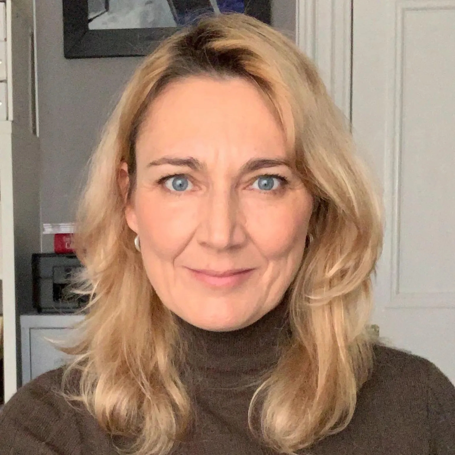 Leasha Esslemont Leasha Esslemont, a woman with shoulder-length, blonde hair wearing a brown turtleneck top. The white door and a painting are visible on the background. 