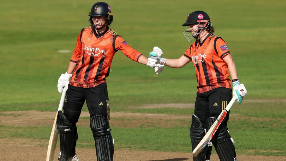 Georgia Elwiss and Kathryn Bryce meet in the middle of the crease to fistpump after a boundary, both are looking onwards. They are both wearing the orange metro bank 2025 Blaze kit.
