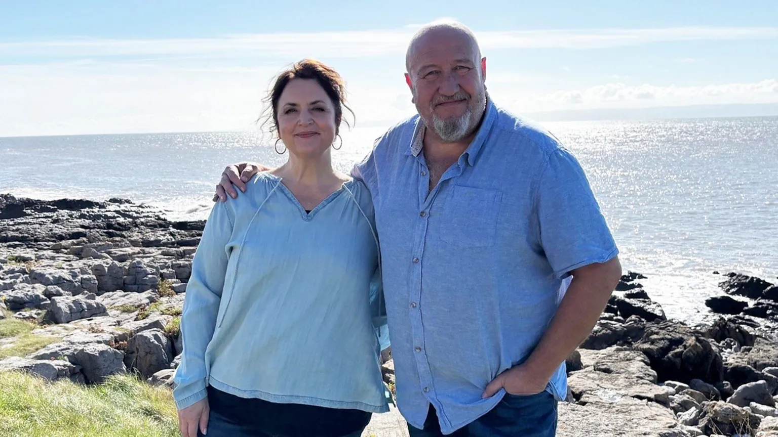 Actors Ruth Jones and Steve Speris stood near a coastline. There are rocks leading the ocean. Ruth is wearing a blue top and Steve has a short sleeved blue shirt on.