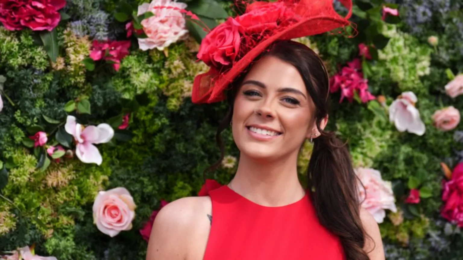  A racegoer stand in front of a wall of flowers. She wears a red dress and red fascinator hat.