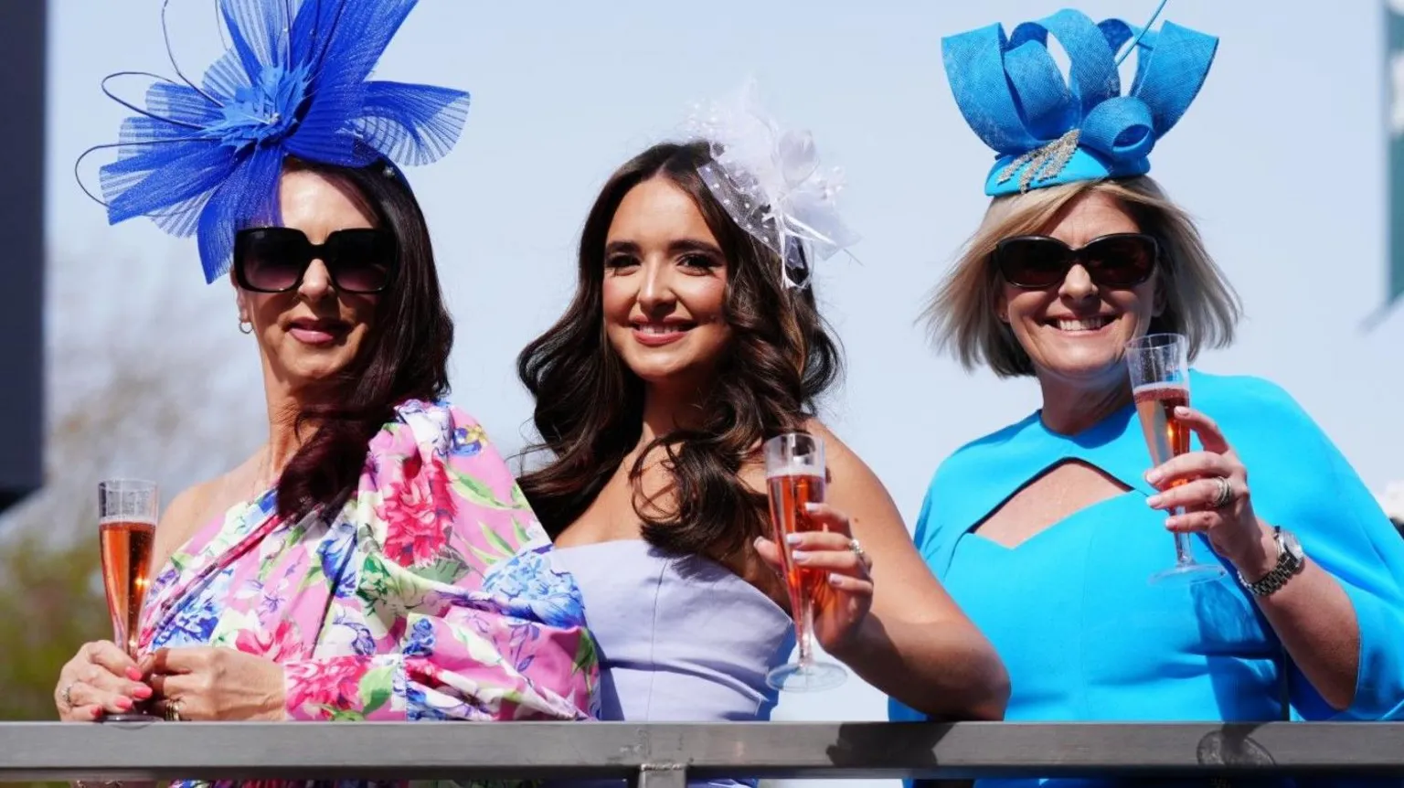  Three women stand along a railing. One is wearing a pink and blue floral dress, and blue fascinator, another is wearing a lilac dress and the third is wearing a blue dress and blue fascinator. They are each holding a glass of pink champagne.