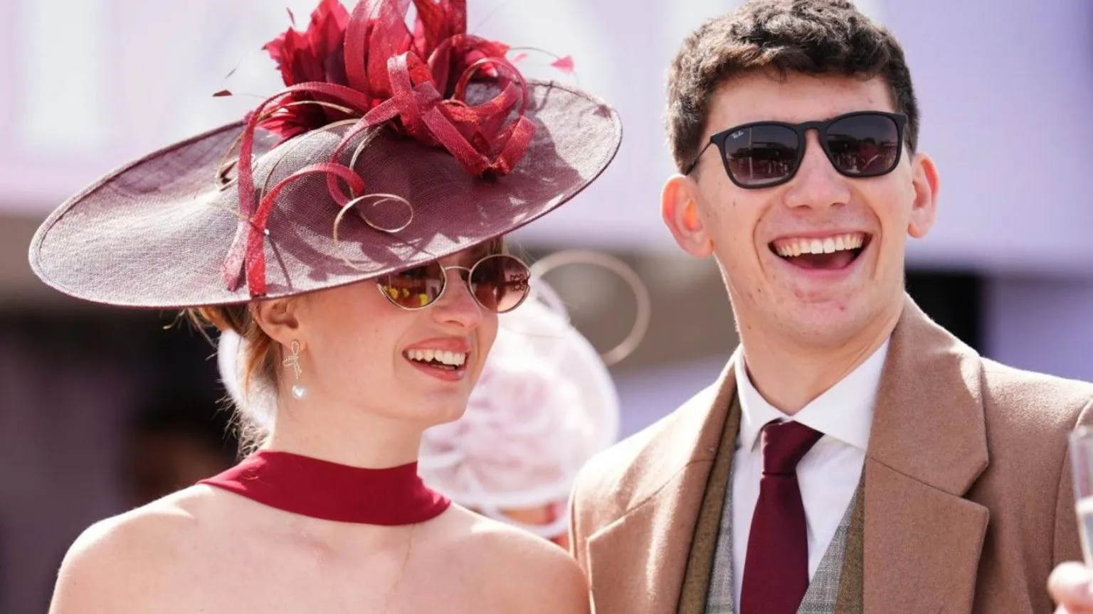  A woman is wearing a dark red dress, fascinator and sunglasses. She is stood with a man who is wearing a tweed suit, beige coat and sunglasses. Both are smiling