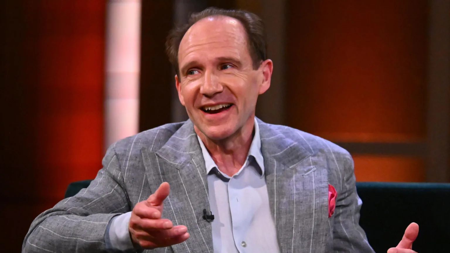  Ralph Fiennes during filming for the Claudia Winkleman Show. He is sitting on a chair and wearing a grey blazer with white pinstripes over a light blue shirt. His hands are rested in conversation. 
