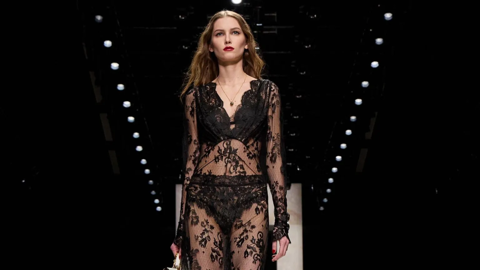  A model walks the runway at the Dolce&Gabbana fashion show during the Milan Fashion Week - Womenswear Fall/Winter 2026/2027 on February 28, 2026 in Milan, Italy. She wears a black lace outfit with long light brown hair and bright red lipstick.