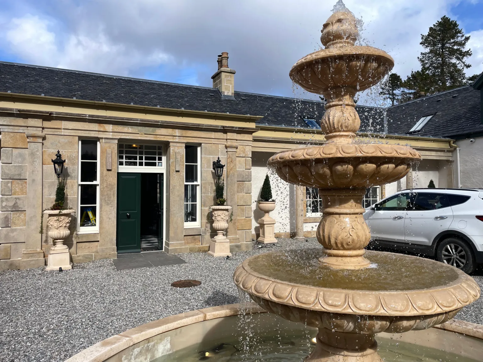 Stone courtyard with a multi-tiered fountain in the foreground, set before a light-coloured house with a green front door, columns, and potted topiary, with a parked car to one side under a partly cloudy sky.