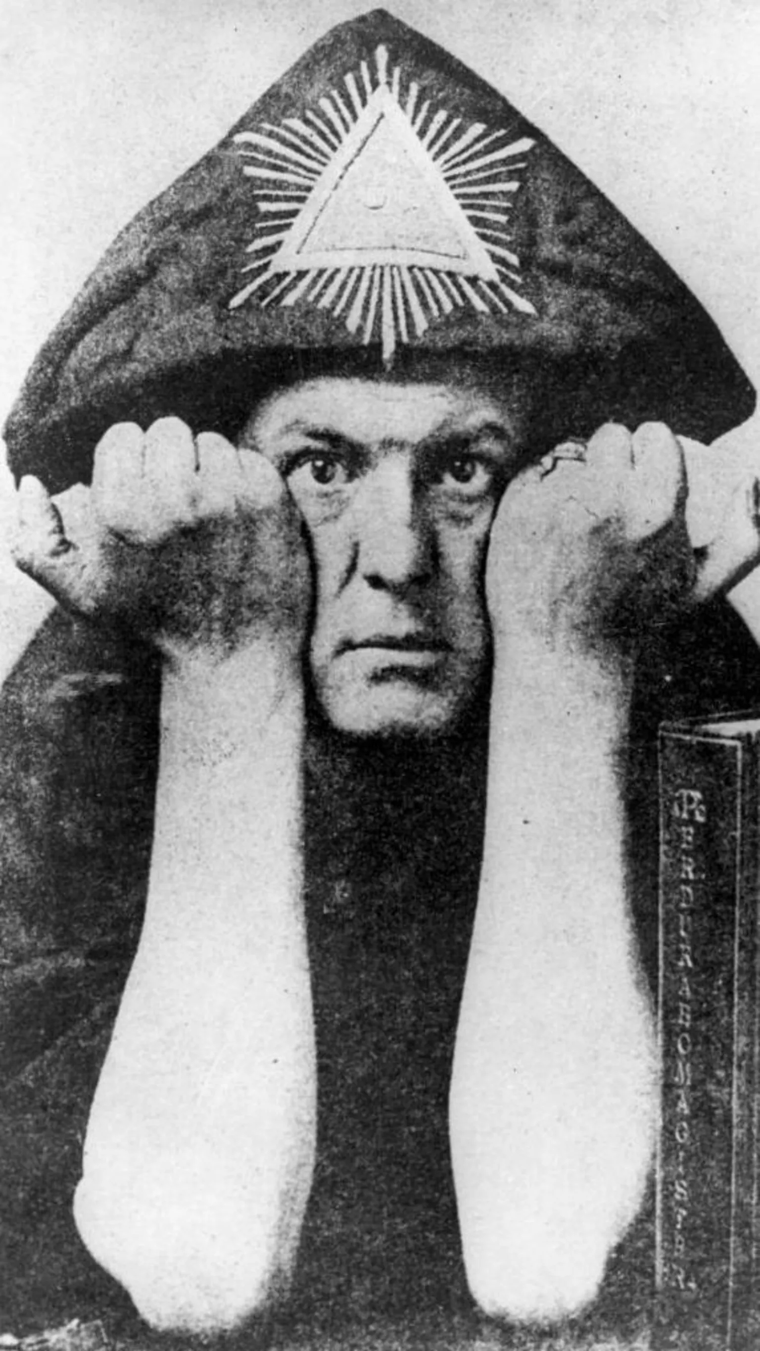  Black-and-white portrait of Aleister Crowley who is seen in a hooded robe marked with a radiant triangle symbol, staring directly at the camera with his fists raised on either side of his face.