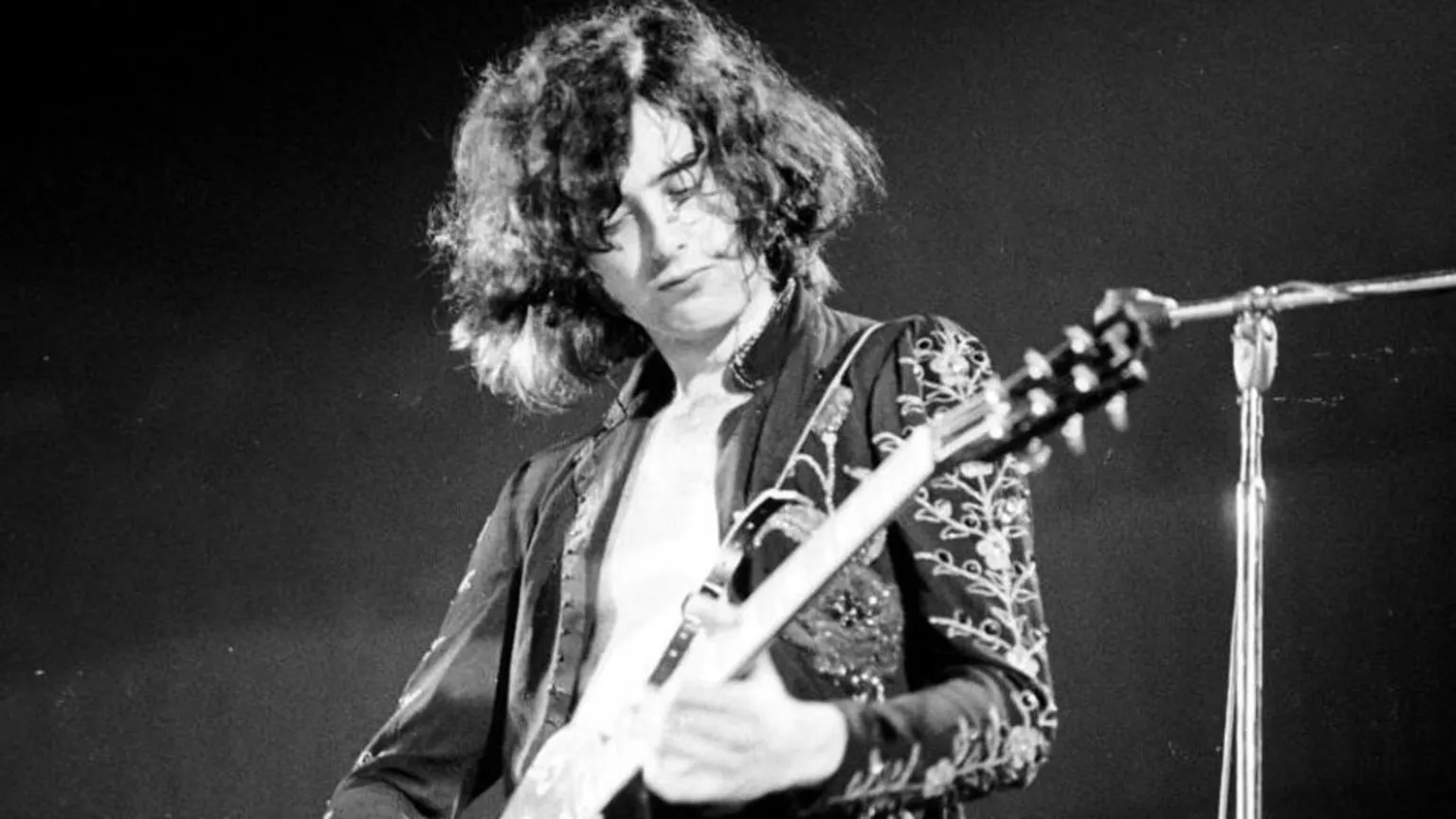  Black-and-white photo of guitarist Jimmy Page, who is seen with long, curly hair playing an electric guitar on stage, wearing an embroidered jacket and looking down in concentration.