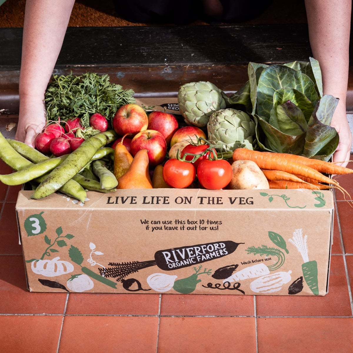 A large vegetable box from Riverford being lifted off a tiled doorstep.