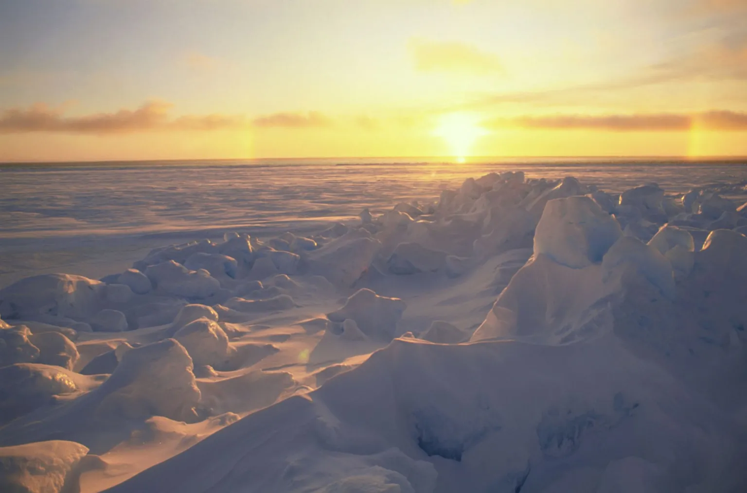  - Doug Allan Sun setting over a frozen sea, illuminating jagged ice formations and snow-covered pressure ridges beneath a golden sky.