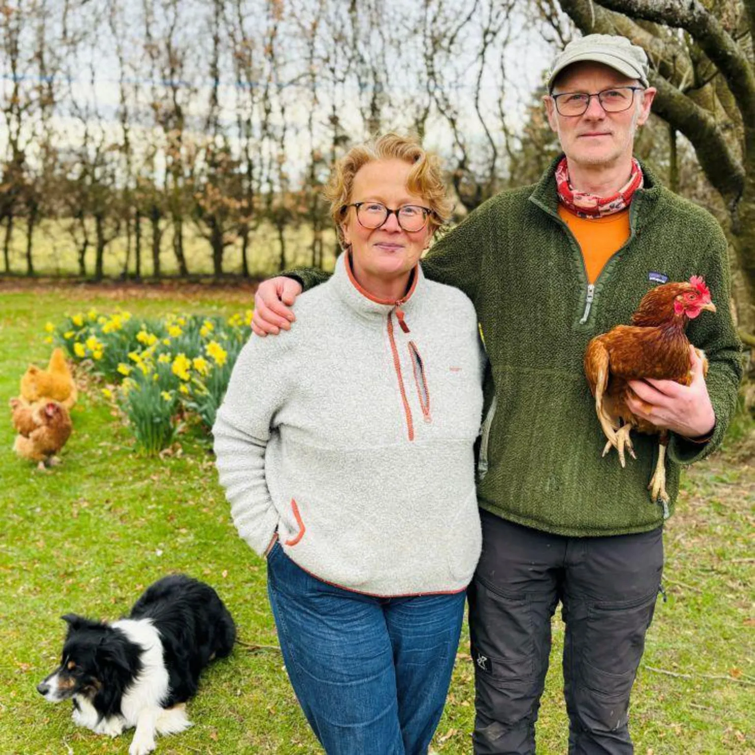 Marguerite Fleming and Frank Richards standing in a garden, he is holding a chicken, there are other chickens in the background, and a black and white collie dog, and daffodils and trees.
