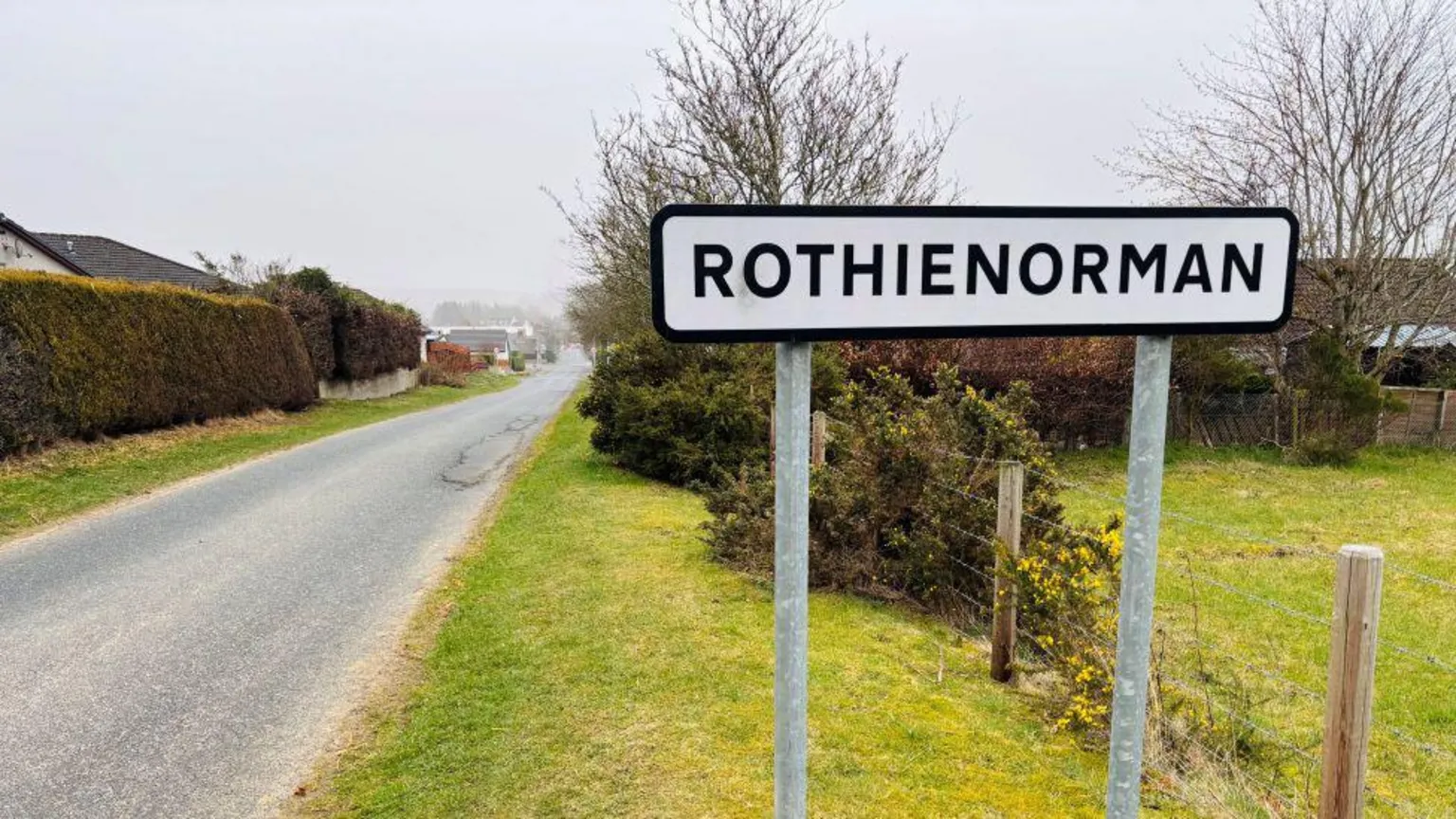 A sign which says Rothienorman, with hedges and homes in the background.