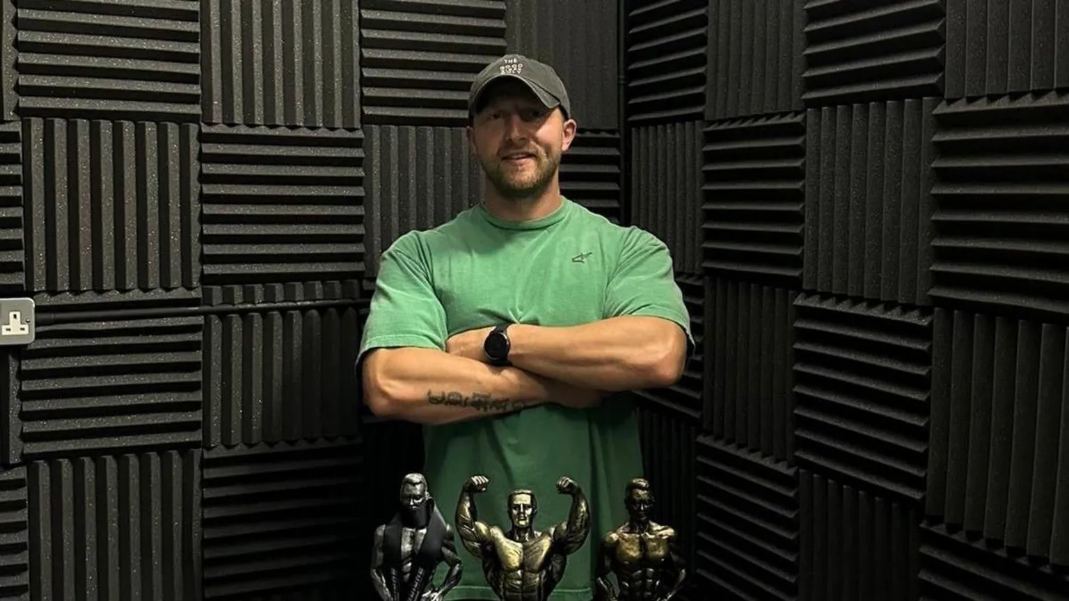 Sion Monty Sion standing with his arms crossed in front of him. He smiles at the camera in front of a black wall. Sion wears a green sports t-shirt and black cap. In front of him are three body building trophies.