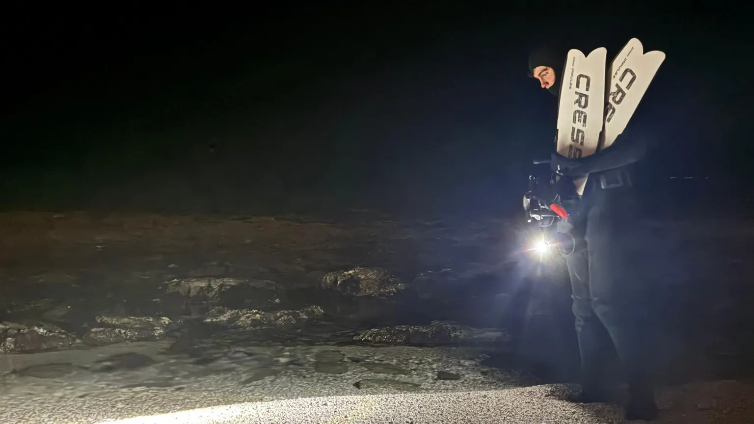Iestyn Morgan Photograph of Iestyn Morgan, pictured stood on the sand. He holds a torch in one hand and flippers in his other hand. He wears a full black wet suit which covers his whole body, bar his eyes and nose. 