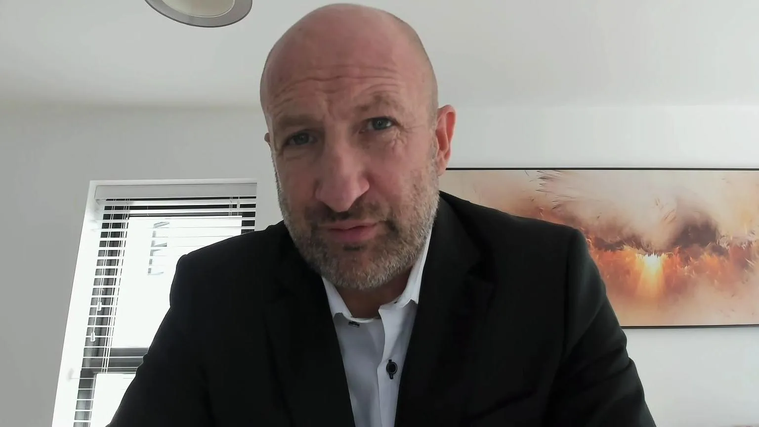 A bald man with a short grey beard, Wearing a white shirt and black blazer, pictured against a white wall with an orange picture in the background.