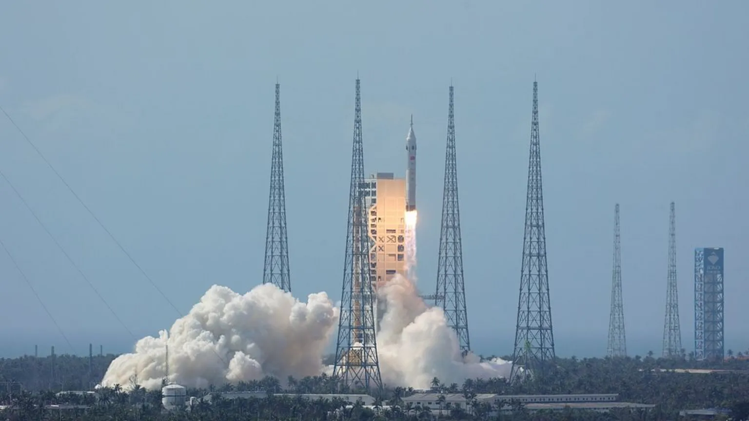 VCG / China Manned Space Agency A dramatic launch photograph taken from ground level shows a large white rocket rising from its launch pad on a column of brilliant orange and white fire. Dense clouds of white smoke and steam billow outward from the base of the rocket across the concrete launch platform, which sits within a tropical coastal landscape — palm trees and a clear blue sky are visible in the background