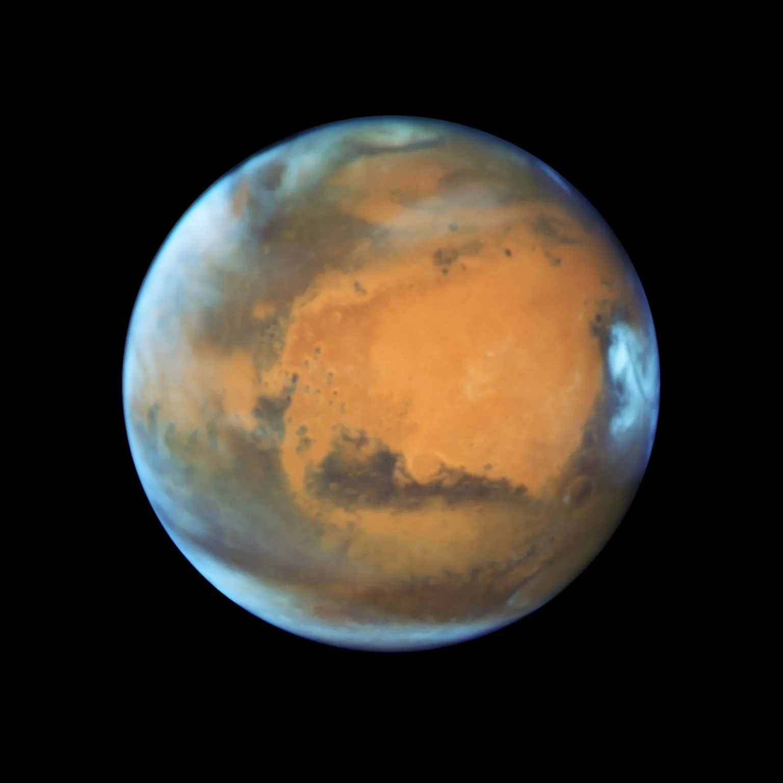 NASA Mars hangs alone against blackness, a glowing, rust‑red marble. The planet’s face is dominated by a vast, soft‑edged orange desert shaped like a sideways Africa. Smoky dark streaks – ancient canyons and volcanic plains – curl around its lower half. Near the top and right edge, bright white clouds and haze cling to the thin atmosphere like torn cotton. A faint bluish rim outlines the curve of the planet, emphasising its roundness and the delicate shell of air wrapped around it.