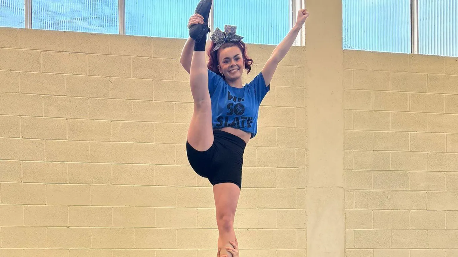 Strikeforce Cheer Clodagh McGonagle is standing holding her leg. She is wearing a blue top and black shorts. And has red hair tied up in a silver bow.