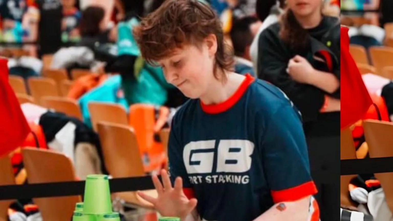 Hsieh Hsin-Ying Laura Beacom has red hair and is stacking green cups on a table. She is looking down at the table and has her hands poised above the cups. She is wearing a dark blue sports top which reads 