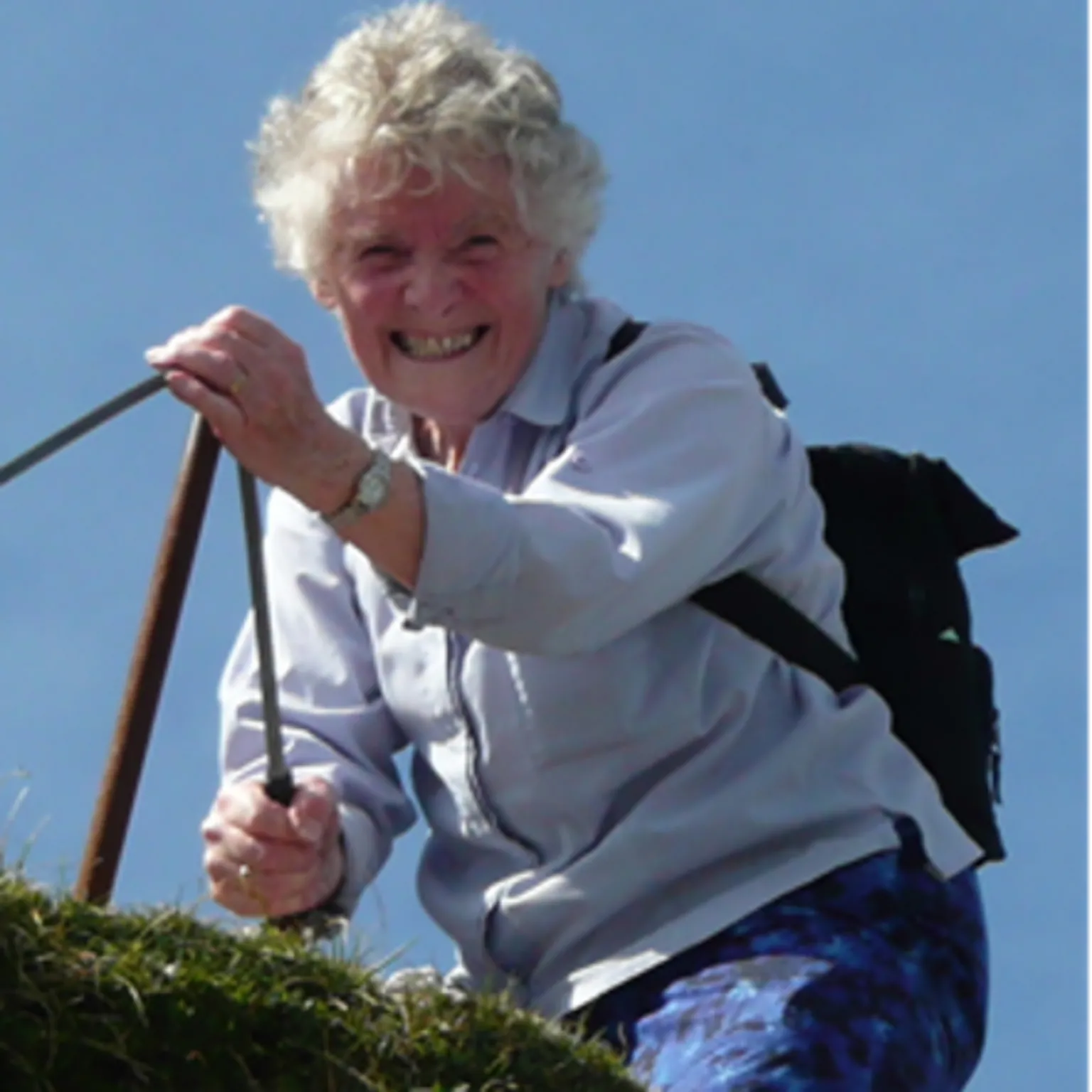 A smiling woman climbing outdoors, holding onto a metal handrail, wearing a light shirt and backpack against a clear blue sky.