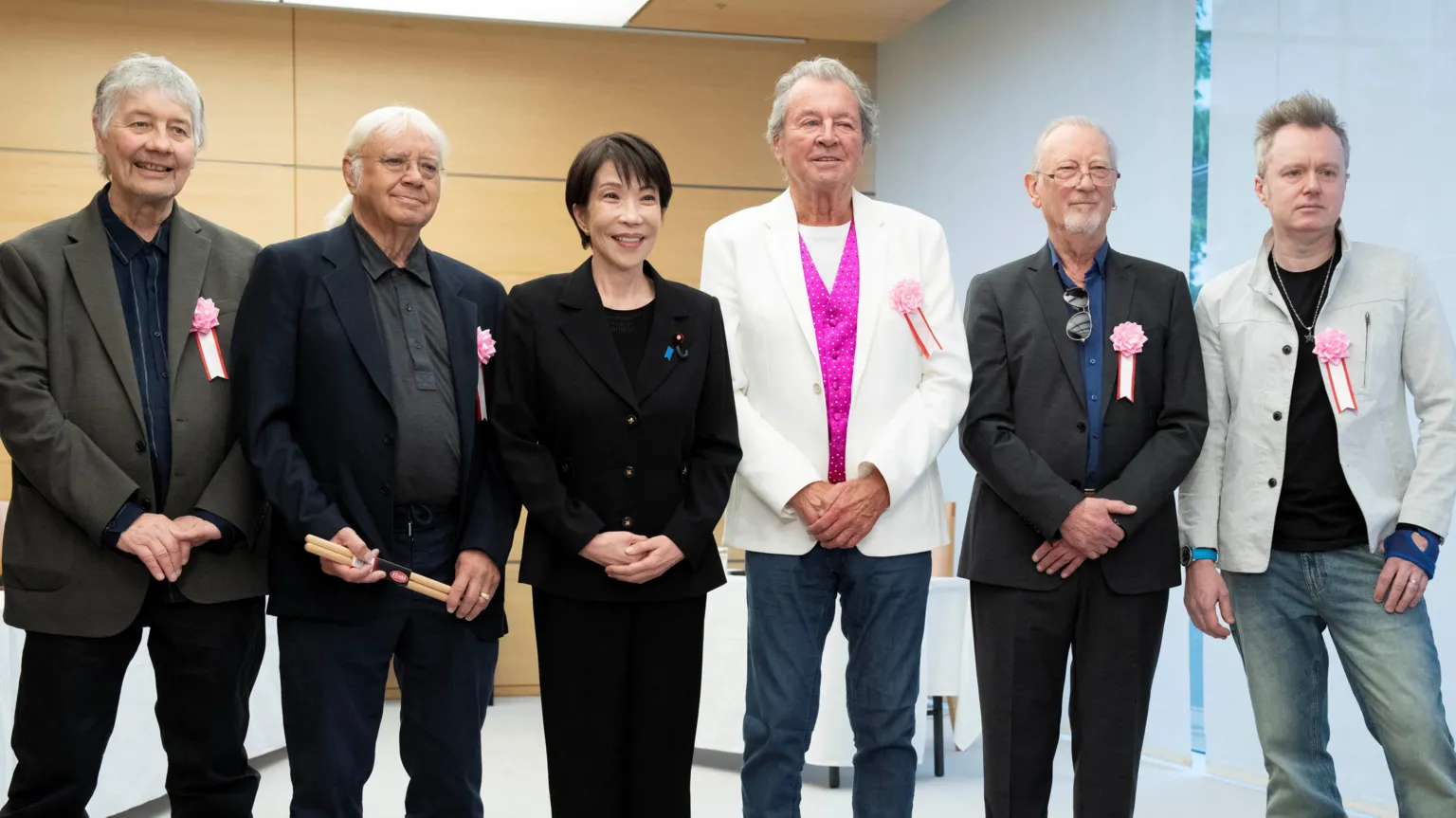 Takaichi poses with Deep Purple. They stand side-by-side in a line and smile. Left to right are: Don Airey, Ian Paice, Sanae Takaichi, Ian Gillan, Roger Glover and Simon McBride. They are stood in a room with wood-panelled walls, and grey blinds drawn over a windowed wall to the right of the frame.