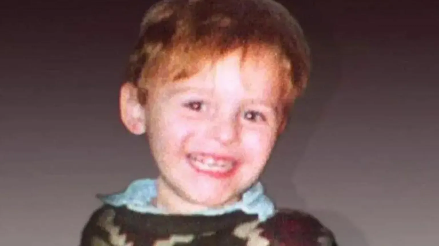  Toddler James Bulger is wearing a brown jumper over a blue shirt and is smiling at the camera.