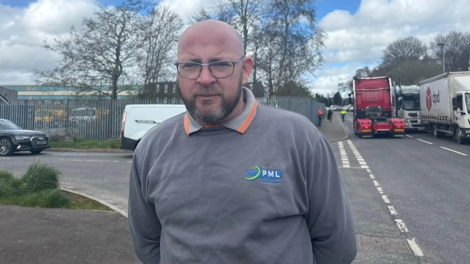 A bald man with a beard and wearing glasses and a grey sweatshirt stands on a road with a number of lorries in the background.