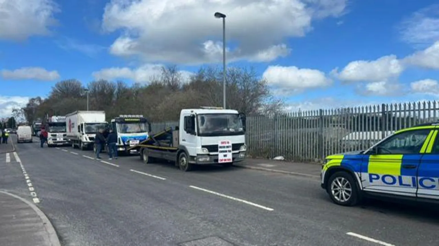 A number of lorries are seen parking on the side of the road in the middle and far ground and closer in the foreground, the side of a Police Service of Northern Ireland jeep can be seen