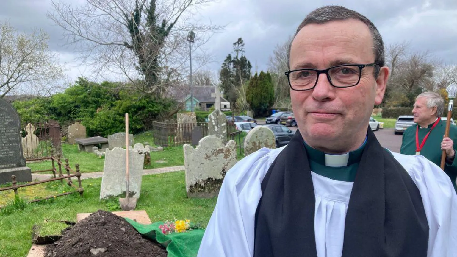 A robed clergy member, a male with short grey hair, wearing glasses stands in the foreground of a graveyard, with an open burial plot covered by green fabric and soil behind them. Gravestones, trees and parked cars are visible in the background, with another robed person standing nearby. 